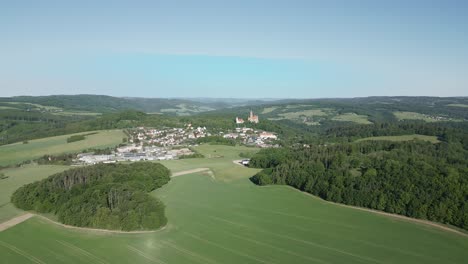 Vista-Aérea-Del-Castillo-Bouzov,-En-Medio-De-Campos-Verdes-Y-Densos-Bosques,-Con-Un-Pequeño-Pueblo-Cercano-Durante-El-Soleado-Día-De-Verano