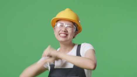 close up of asian woman worker wearing goggles and safety helmet singing and dancing while standing in the green screen background studio