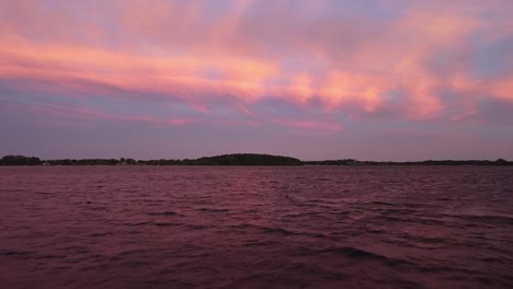 Aerial-view-of-drone-flying-just-over-the-surface-of-the-water-at-sunset-showing-pink-clouds-and-smaller-white-clouds-with-a-forest-in-the-background