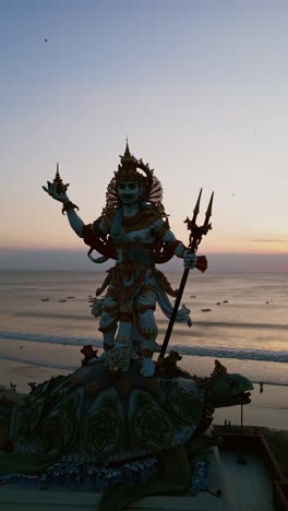 the baruna statue stands majestically above kuta beach while golden sunset light illuminates the coastline, captured in stunning vertical aerial footage highlighting both statue and ocean horizon