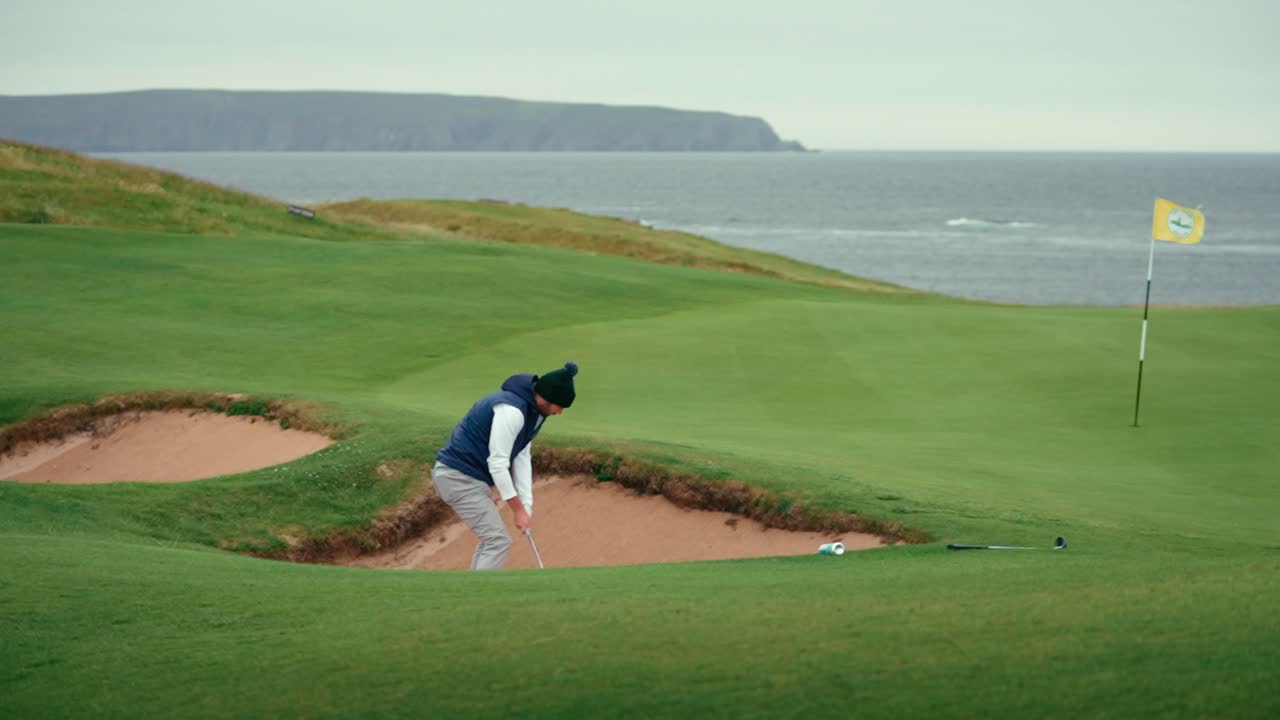 Male golfer hits ball out of pot bunker hazard with sand wedge to land ball near hole and flag stick on coastal Ireland golf course green