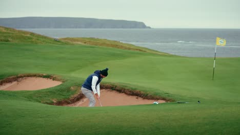 male golfer hits ball out of pot bunker hazard with sand wedge to land ball near hole and flag stick on coastal ireland golf course green