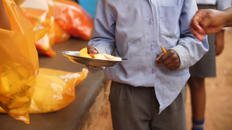 Teachers of rural African school placing fresh pieces of fruit in young pupil's plate for lunch