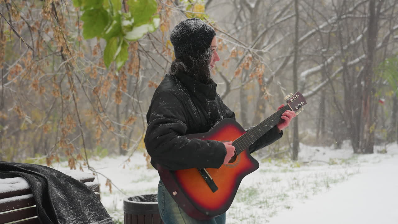 vista lateral de una mujer con capucha negra tocando guitarra acústica al aire libre en un parque nevado, rodeada de árboles desnudos helados y nieve que cae suavemente, creando un momento de invierno sereno lleno de música