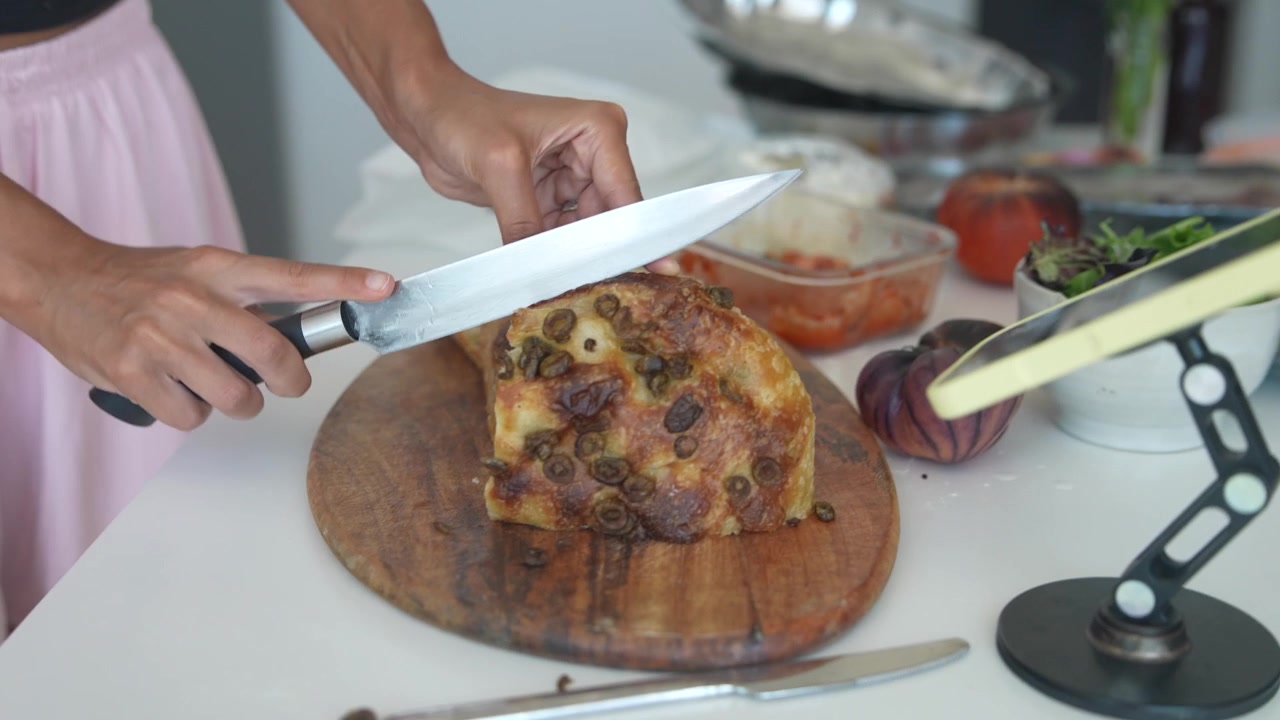 Mujer rebanando pan focaccia fresco de aceitunas en tabla de madera.
