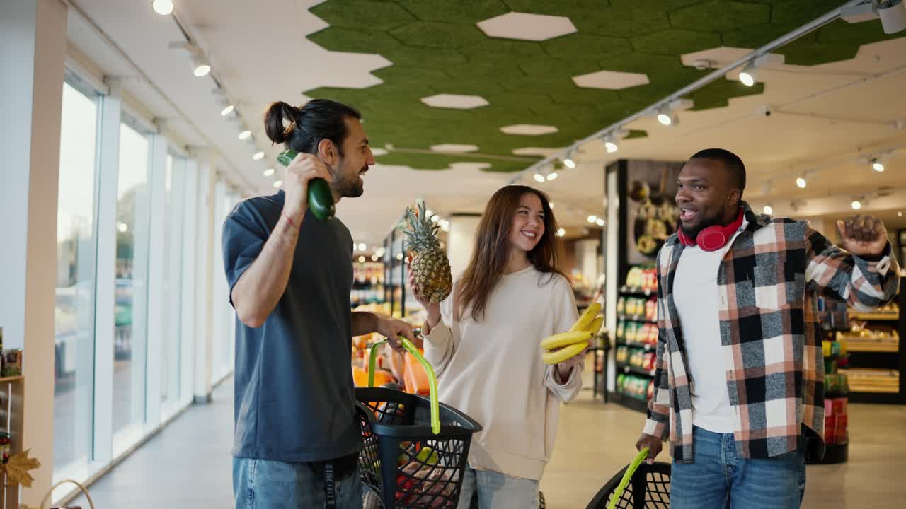 색 셔츠와 빨간색 무선 헤드폰을 입은 검은 피부를 가진 남자가 그의 친구들과 함께 회색 티셔츠를 입은 소녀와 갈색 남자가 현대 식료품 슈퍼마켓의 카운터 근처에 다양한 과일과 채소를 손에 들고 춤을 추고 있습니다.