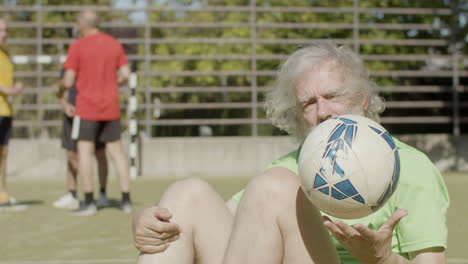 sonriente jugador de fútbol senior sentado en el campo, lanzando la pelota con la mano y mirando a la cámara