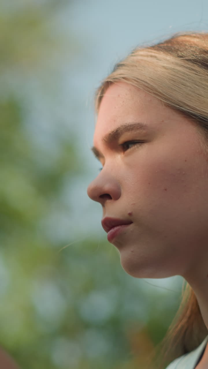 Thoughtful young woman outdoors with sunlight softly illuminating her face, adjusting her hair back pinned with black pin, wearing cyan top, under blurred sky, with blurred view of green tree