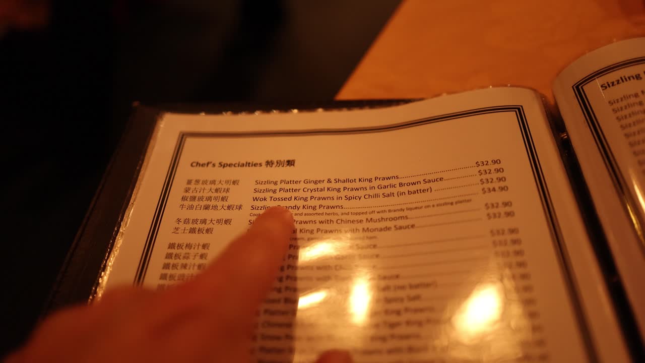 Person’s hand points and flips through laminated Chinese menu under warm indoor lighting, close-up