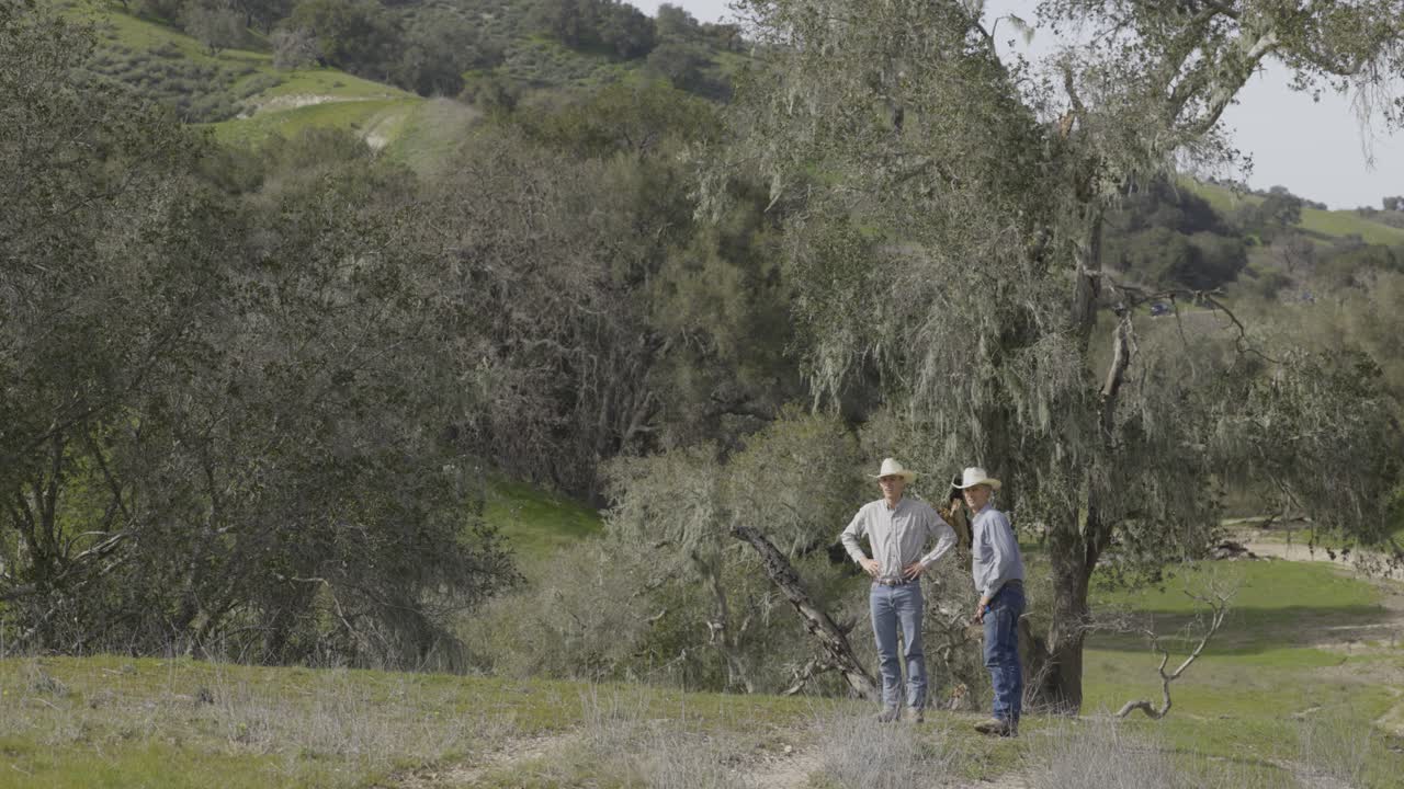 Two cowboys standing in a rural landscape with trees and hills