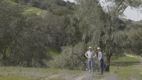 two cowboys standing in a rural landscape with trees and hills