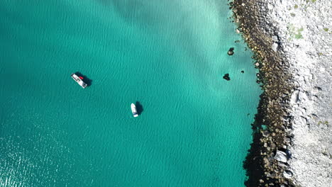 Downward-angle-drone-footage-of-two-boats-in-clear-blue-waters-at-the-island-of-Vaeroy,-Lofoten-Islands-in-Norway