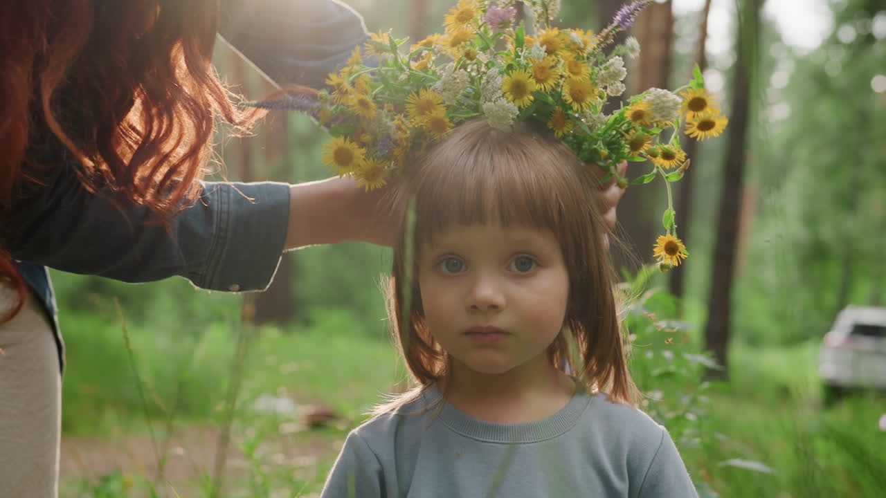 Portrait of schoolgirl standing in forest as mother decorates her head with colorful wildflower bouquet, surrounded by green nature, sunlight, and warm atmosphere showing tenderness, and family love