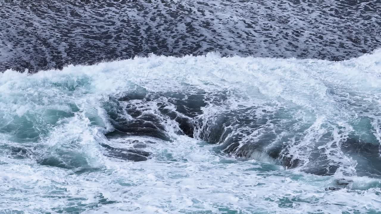 Powerful ocean waves crash against dark rocks, foaming and swirling under overcast natural daylight