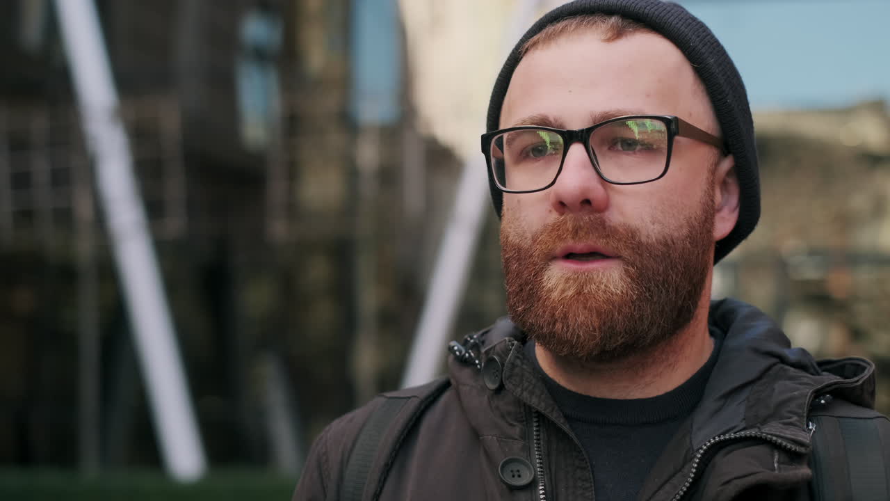 Close-up portrait of a man with a beard, glasses, and beanie in an urban setting