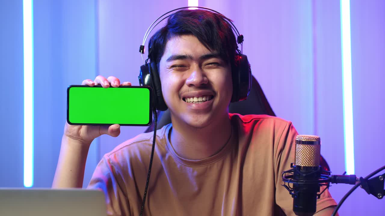 Uomo asiatico che tiene in mano un telefono cellulare con uno schermo verde di mockup mentre è in diretta streaming.