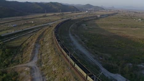 full loaded coal cargo carriages train stopped on a long turn of a railway in green fields, with road and mountain ridge in the background, on the sunset