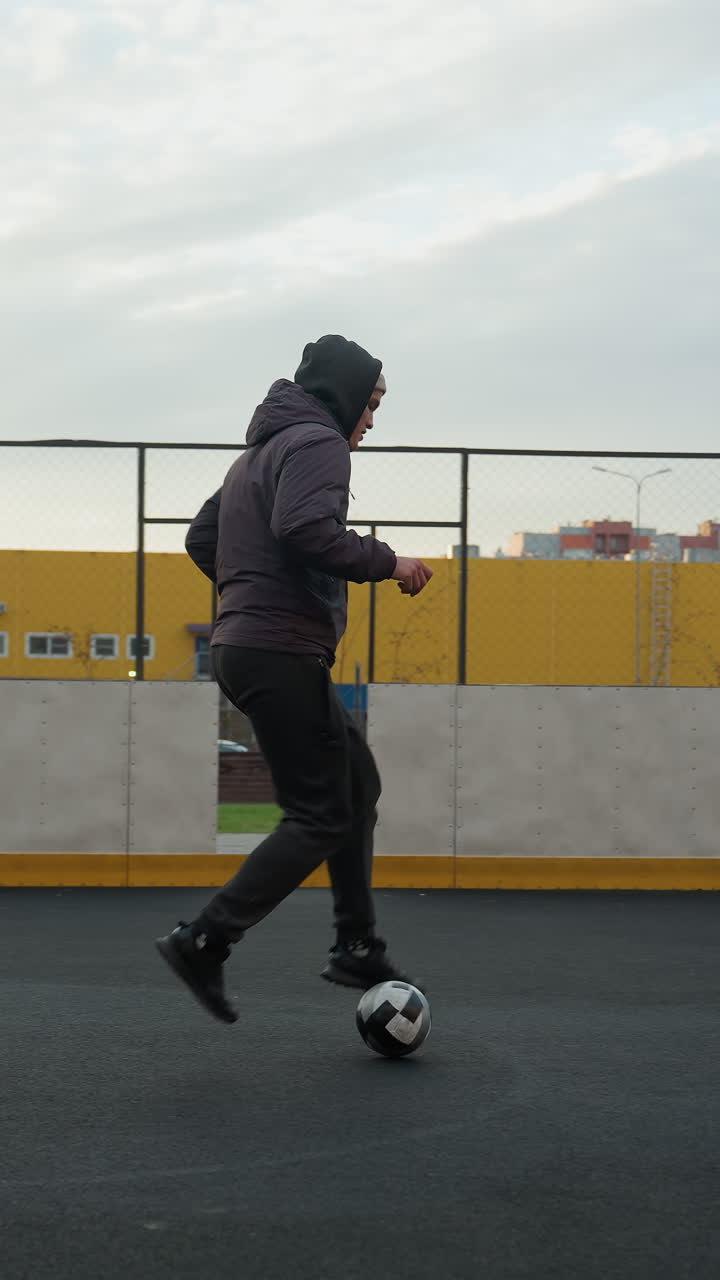 Athlete training with football on sport arena tapping ball forward with precision, showcasing control and agility, background features urban buildings, yellow wall, people walking, and fencing