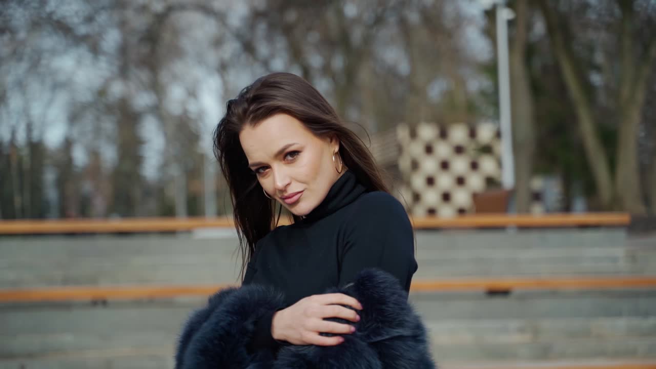 Portrait of beautiful model. Glamour girl in black fur coat posing on camera outdoors. Attractive brunette in the park. Camera moves around.