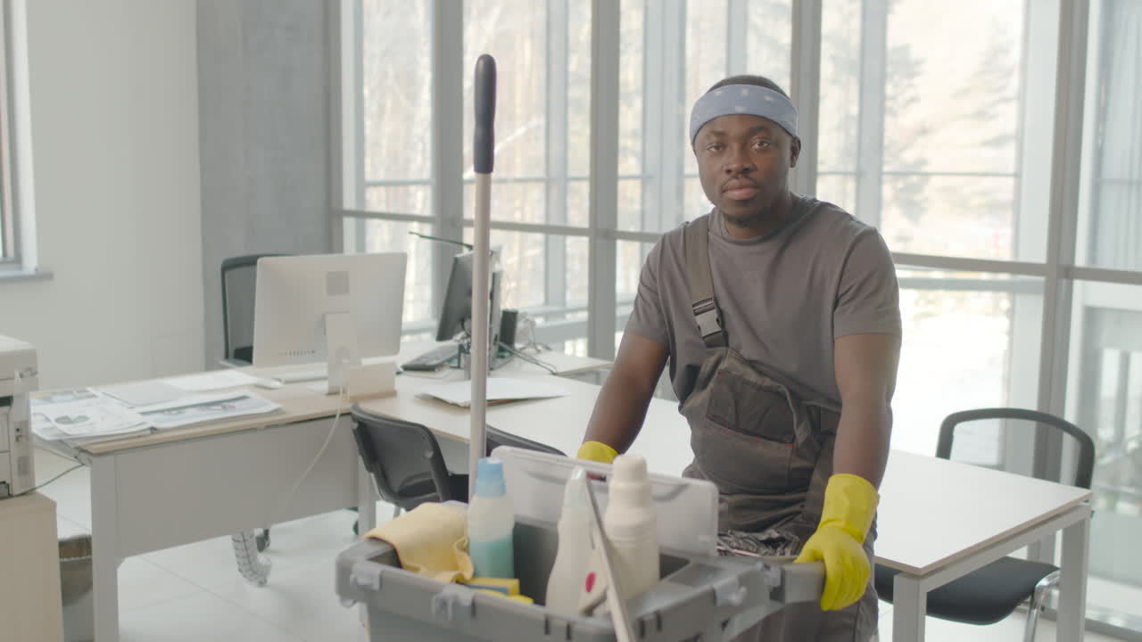 Cleaning Man Wearing Gloves And Bandana Posing With Cleaning Cart And Looking At The Camera Inside An Office
