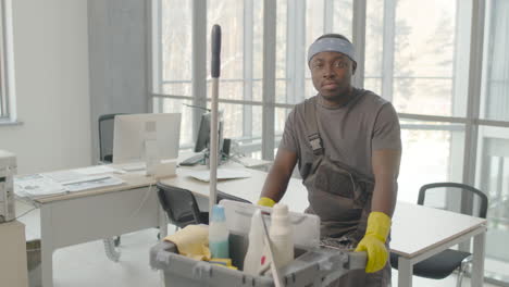 cleaning man wearing gloves and bandana posing with cleaning cart and looking at the camera inside an office