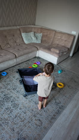 small male kid stands in the messy room with a suitcase in the middle. toddler picks his clothes from the floor. black cat comes up to the boy. high angle view. vertical video.