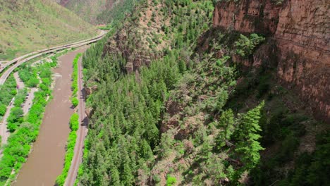 aerial shot flying down the face off rocky canyon gorge cliffside covered in healthy green alpine trees and foliage during bright hot warm summer day near colorado river. glenwood canyon colorado usa