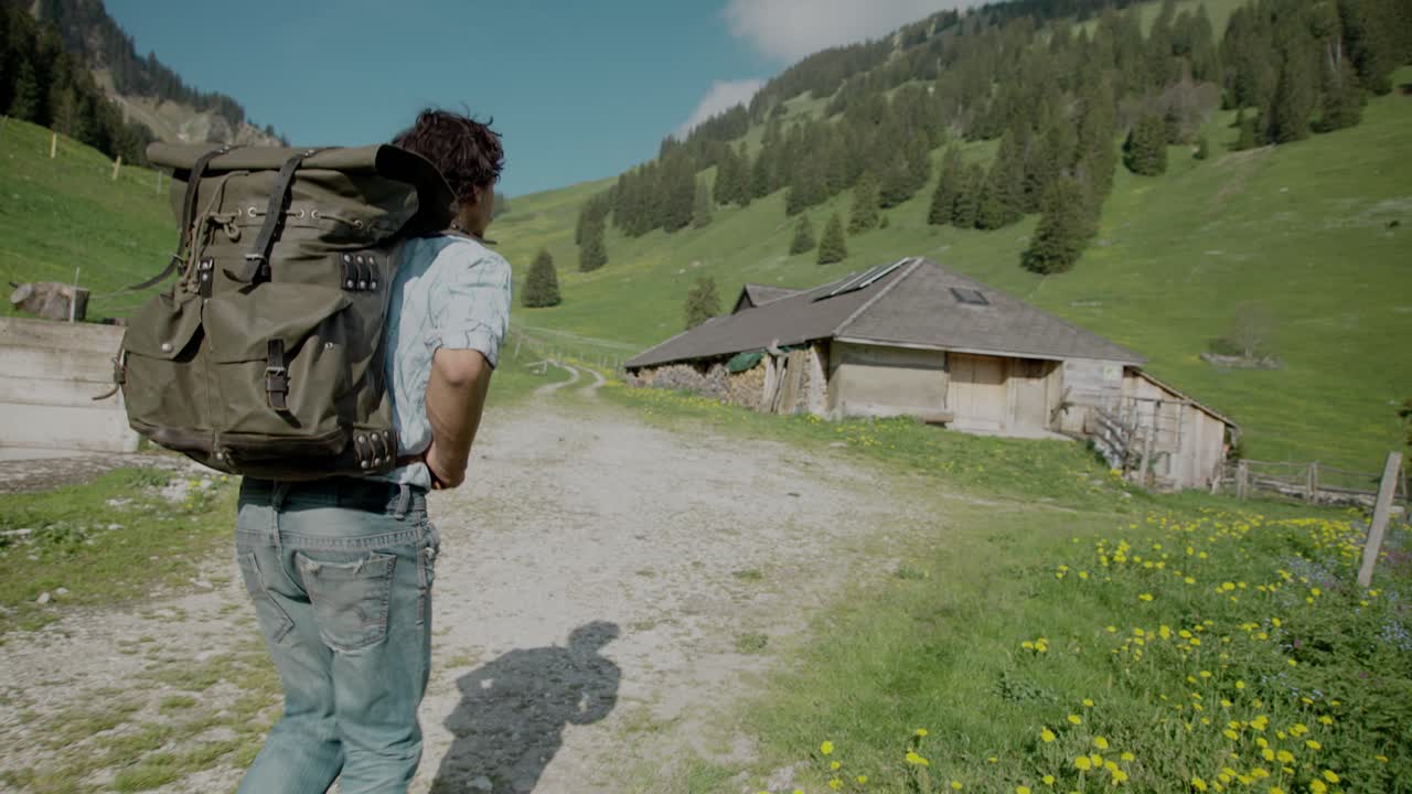 Gimbal tracking backpacker in casual clothing hiking on rural dirt road in mountain valley trail towards a house