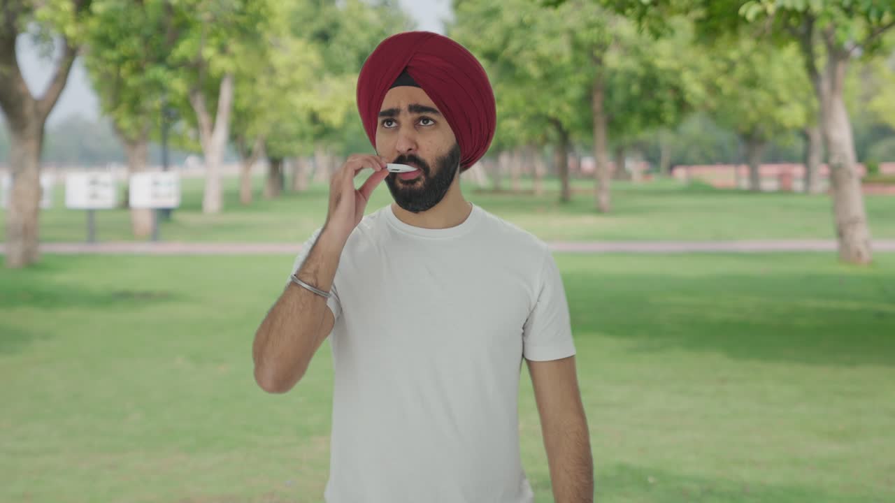 Uomo indiano Sikh malato usa un termometro per controllare la febbre al parco.