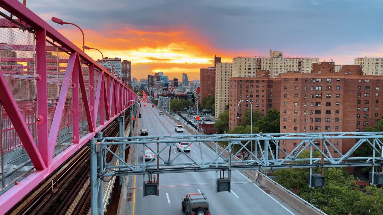 New York, USA, 2 marzo 2026: Prospettiva della passerella del Ponte di Williamsburg al tramonto con vista sulla città. Inquadratura simmetrica dei tralicci rosa del ponte e dell'autostrada che conduce verso un brillante tramonto arancione