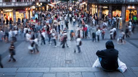 a bustling street filled with crowds during a weekend evening. people are walking past shops while one individual sits calmly on a step, surrounded by the city's vibrant energy.