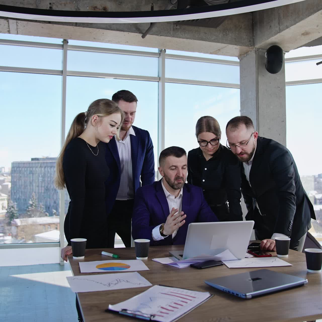 Teammates gathered around their boss to work out solutions. Chief sitting at the laptop and employees standing around and suggesting ideas