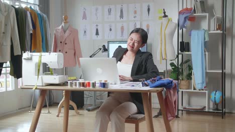 diseñadora asiática en traje de negocios trabajando en una computadora portátil y teniendo dolor de espalda mientras diseña ropa en el estudio