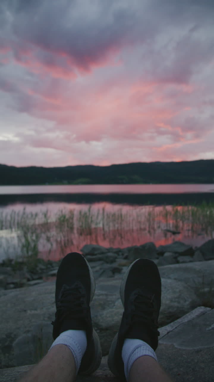 Vertical view of a persons feet, he sits by a lake with feet on rocks, looking at a pink and purple sunset reflecting on calm water.