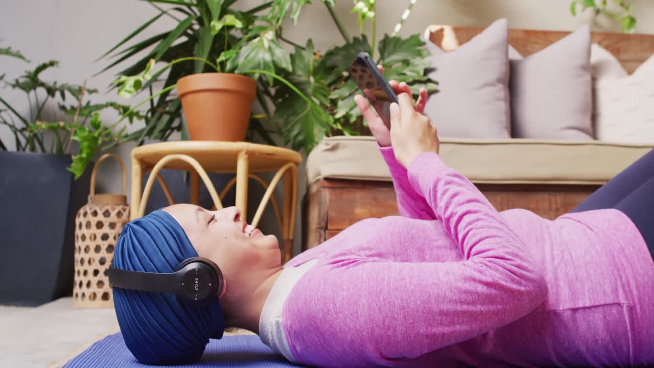 video de una mujer biracial sonriente con hijab y auriculares acostada en una alfombra de yoga usando un teléfono inteligente.