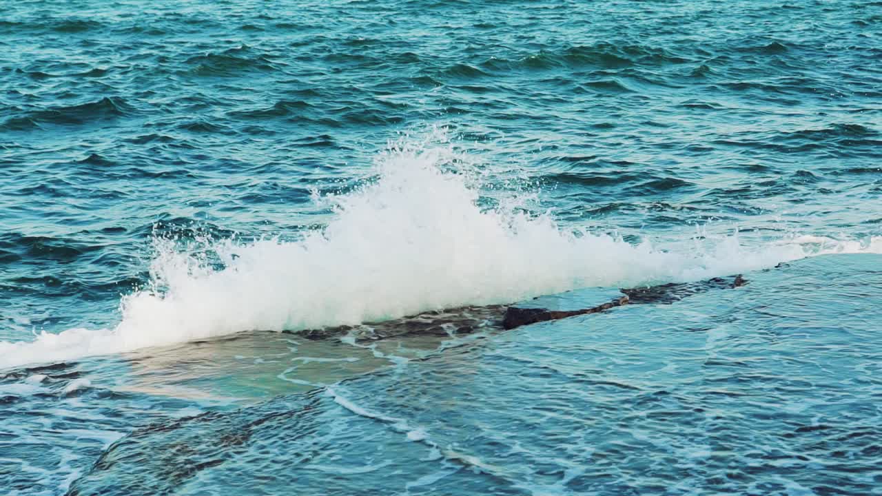 Turquoise waves is colliding with stone rocks near the shore and turning into thousands of drops of water. Slow motion. View from above