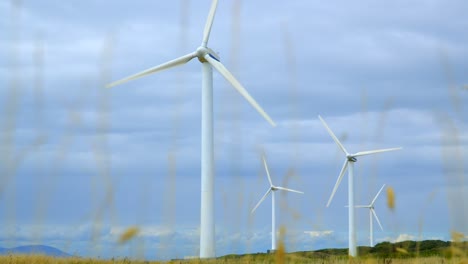 Windturbinen-Und-Heller-Bewölkter-Himmel-Mit-Vergilbtem-Gras-Im-Vordergrund,-Das-Sich-Mit-Zunehmendem-Aufstieg-Auflöst