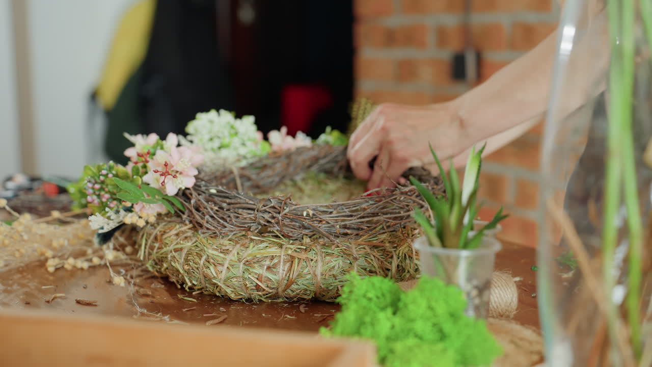 Close up of hands arranging pink flowers and greenery on rustic wreath base made of straw and twigs, showing detailed floral decoration process, emphasizing creativity, craft, and handmade natural decor