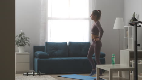 Una-Mujer-Deportiva-Relajante-Está-Bebiendo-Agua-Durante-El-Descanso-En-Casa,-Entrenando-De-Pie-En-La-Sala-De-Estar-En-Un-Día-Soleado,-Bienestar-Y-Fitness