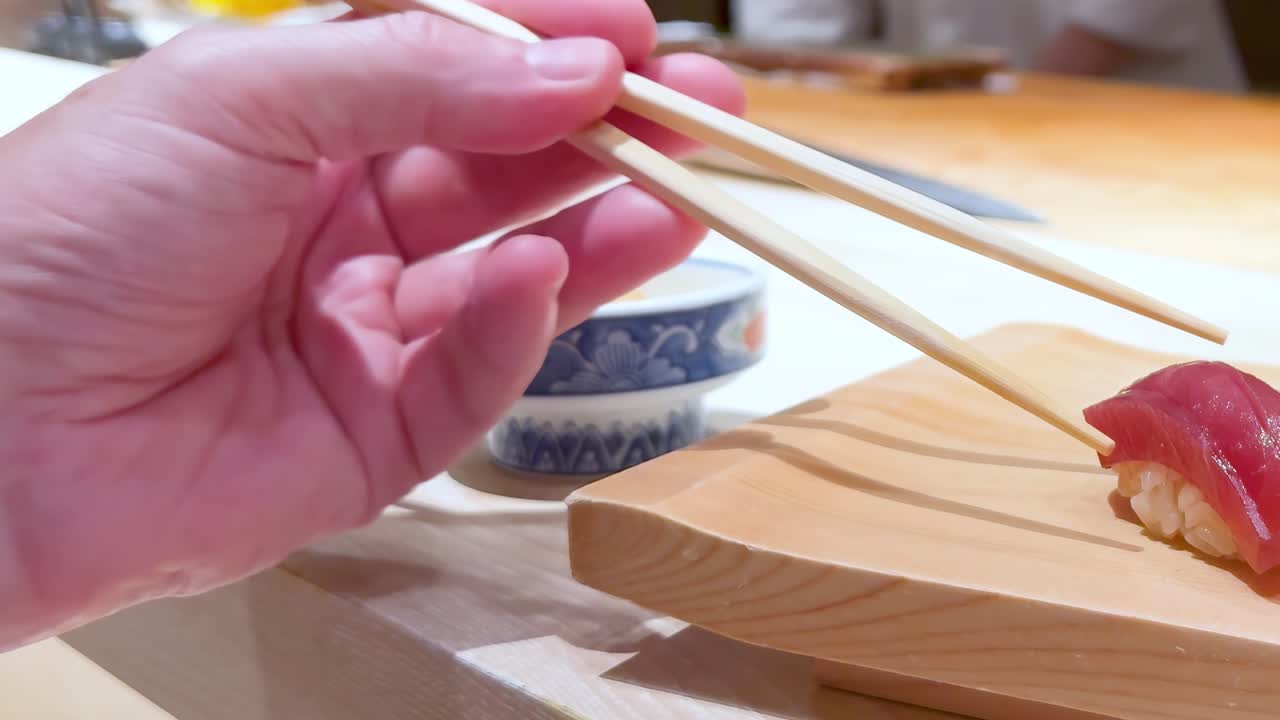 Close-up of chopsticks picking up sushi from a wooden board with a small bowl nearby.