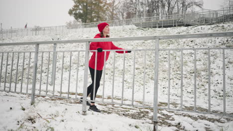 woman exercising outdoors during winter, holding iron railing while stretching her leg outward, wearing black gloves, with foggy atmosphere, snowy ground, and distant trees in the background
