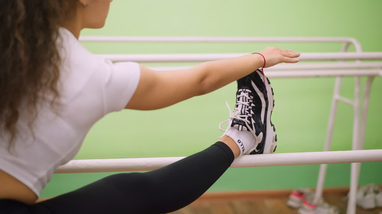 Side view of girl in white top and black leggings stretching leg on white bars indoors, black sneakers with white socks visible, arm extended forward for balance, exercise showing strength, focus