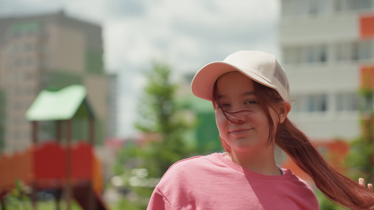 Girl In Cap Smiling Outdoors Near Colorful Apartments, Posing Confidently And Twirling Braid In Soft Sunlight, Casual Sweatshirt And Relaxed Summer Mood, Candid Closeup With Playful Expression