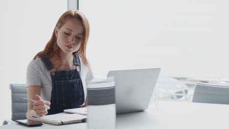 Joven-Empresaria-Vestida-Informalmente-Trabajando-En-Una-Computadora-Portátil-En-Una-Oficina-Moderna-Atendiendo-Una-Llamada-Por-Teléfono-Móvil