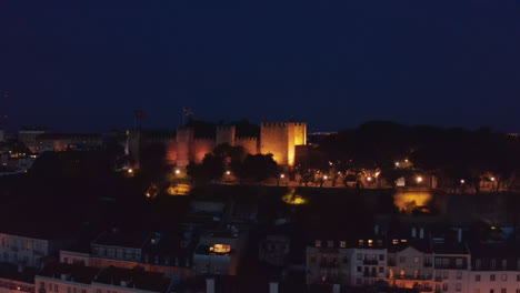 Night-aerial-view-of-old-Saint-George-Castle-on-hill-above-town.-Illuminated-walls-and-defence-towers.-Drone-flying-towards-medieval-landmark.-Lisbon,-capital-of-Portugal.