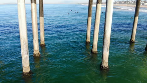 cool, awesome, long aerial 4k drone lateral tracking fly by of huntington beach pier pilings, past paddling surfer in waves, boom up to reveal lifeguard tower - pier, and full beach - downtown area