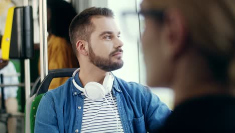 Portrait-Of-The-Young-Handsome-Guy-With-Big-White-Headphones-On-His-Neck-Sitting-In-The-Tram-And-Looking-In-The-Window,-Then-Turning-Face-To-The-Camera-And-Smiling-Cheerfully-To-The-Camera