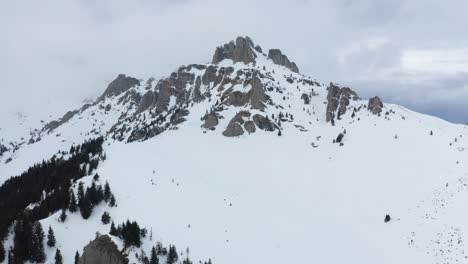 Snow-capped-Ciucas-Mountains-under-overcast-sky,-pine-trees-in-foreground,-aerial-view