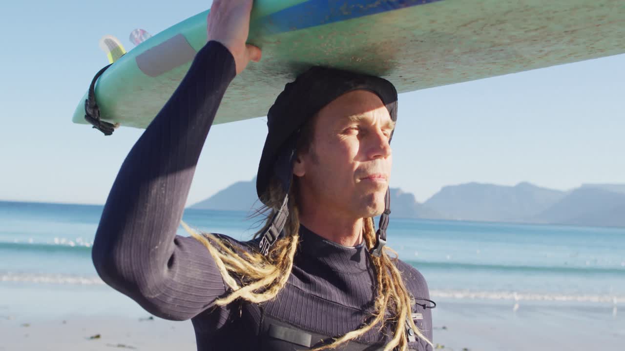 video de un hombre caucásico sonriente con rastas llevando una tabla de surf en la cabeza en una playa soleada
