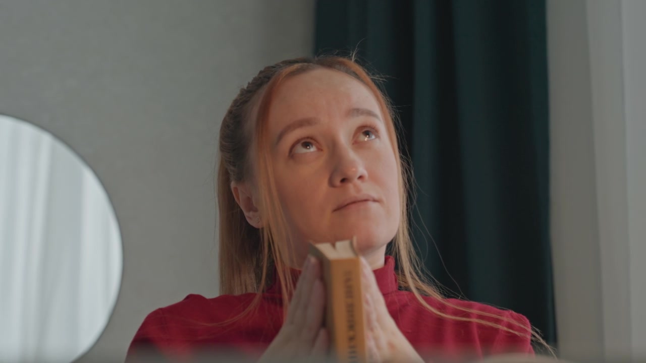 Jovem mulher com cabelos longos presos lê um livro intitulado. A mulher veste uma blusa vermelha enquanto segura o livro com as duas mãos. Espelho redondo em uma parede clara e um objeto retangular vertical.